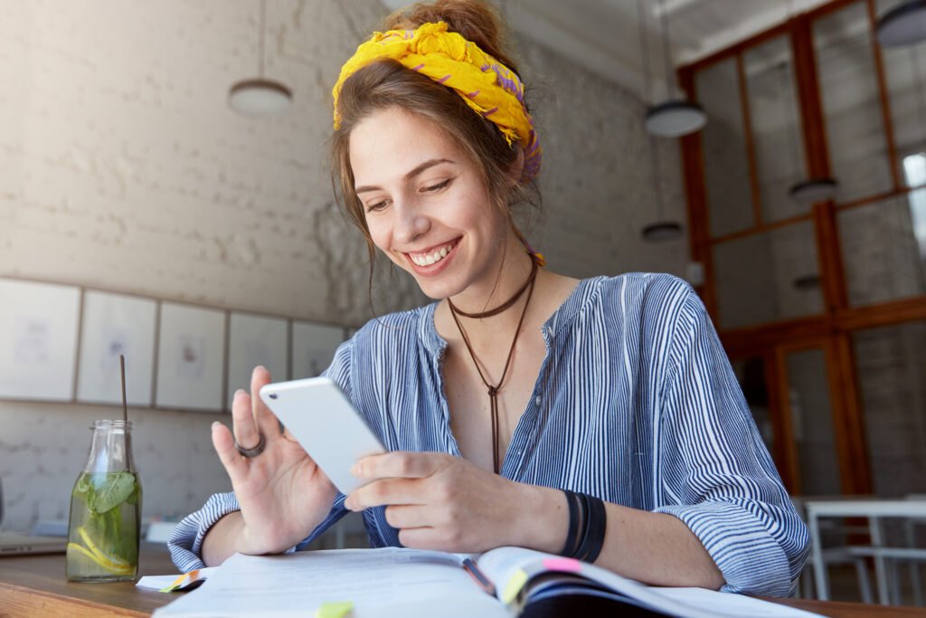 happy student girl with charming smile reading positive news or browsing newsfeed via social networks using mobile phone, having small break while doing homework, sitting at cafe and drinking lemonade