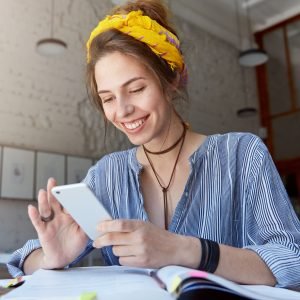 happy student girl with charming smile reading positive news or browsing newsfeed via social networks using mobile phone, having small break while doing homework, sitting at cafe and drinking lemonade