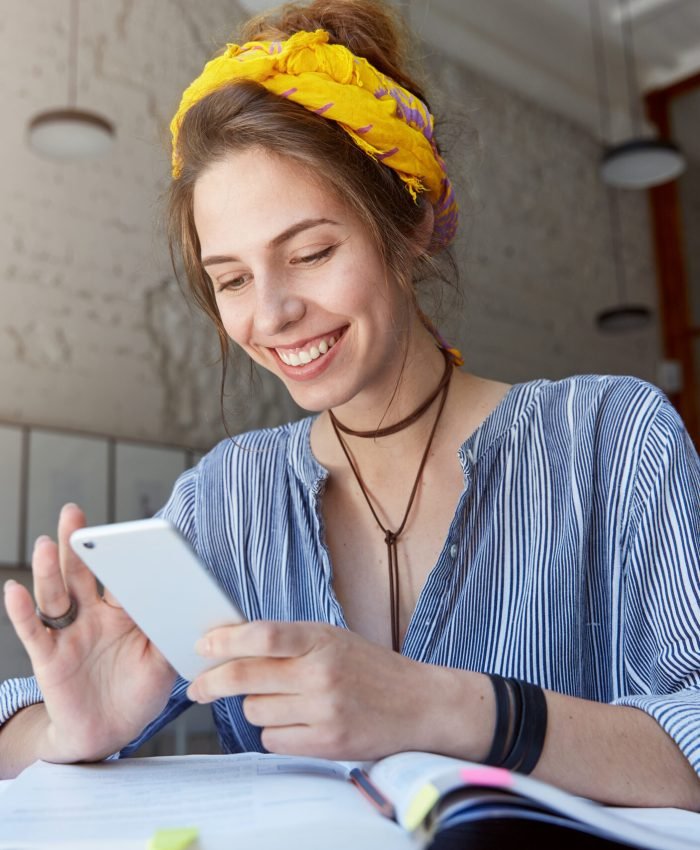 happy student girl with charming smile reading positive news or browsing newsfeed via social networks using mobile phone, having small break while doing homework, sitting at cafe and drinking lemonade